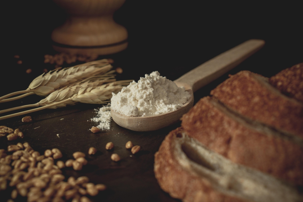 Uma colher de farinha de trigo, grãos de trigo ao lado da colher e um pão de centeio colocado do outro lado, todos em cima de uma mesa de madeira.